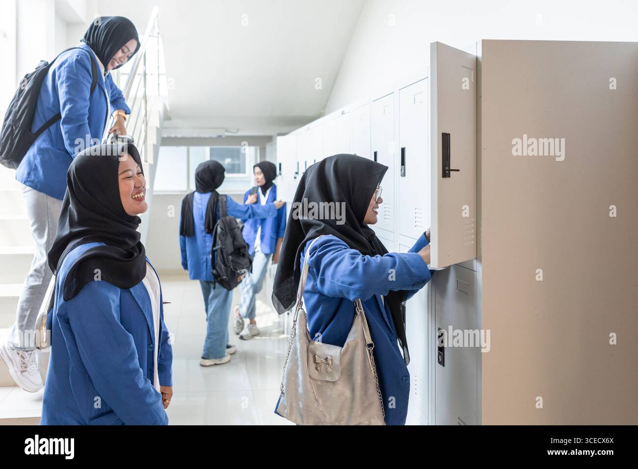 Asiatisch-muslimische Schüler treffen sich in einem hellen Flur, um sich auf einen neuen Schultag vorzubereiten. Sie interagieren mit ihren Schließfächern und untereinander und fördern so eine bestellung Stockfoto