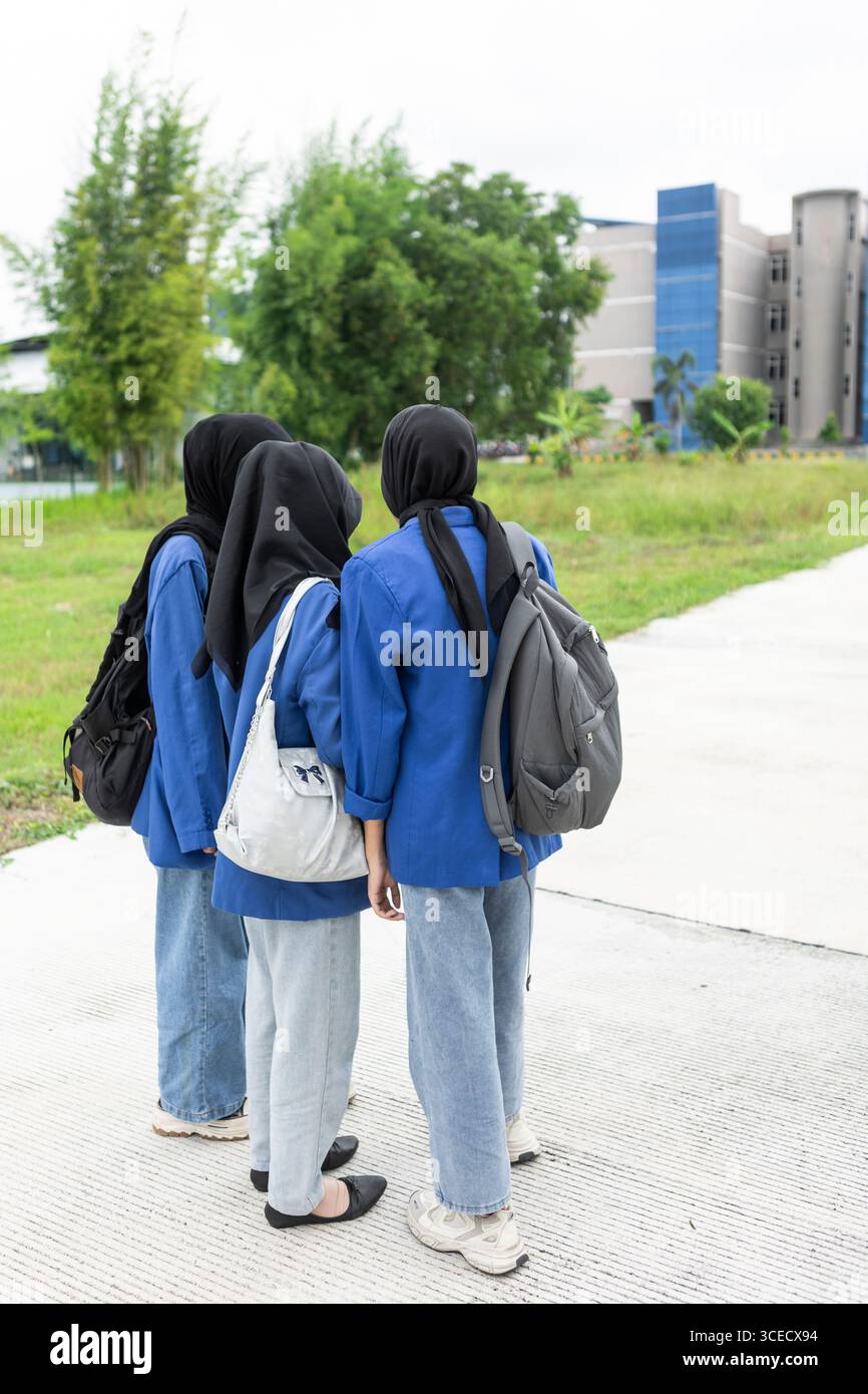 Drei asiatische muslimische Frauen in blauen Uniformen stehen mit Rucksäcken zusammen und bereiten sich auf ein neues akademisches Jahr vor. Die Outdoor-Umgebung unterstreicht ihre Schwärme Stockfoto