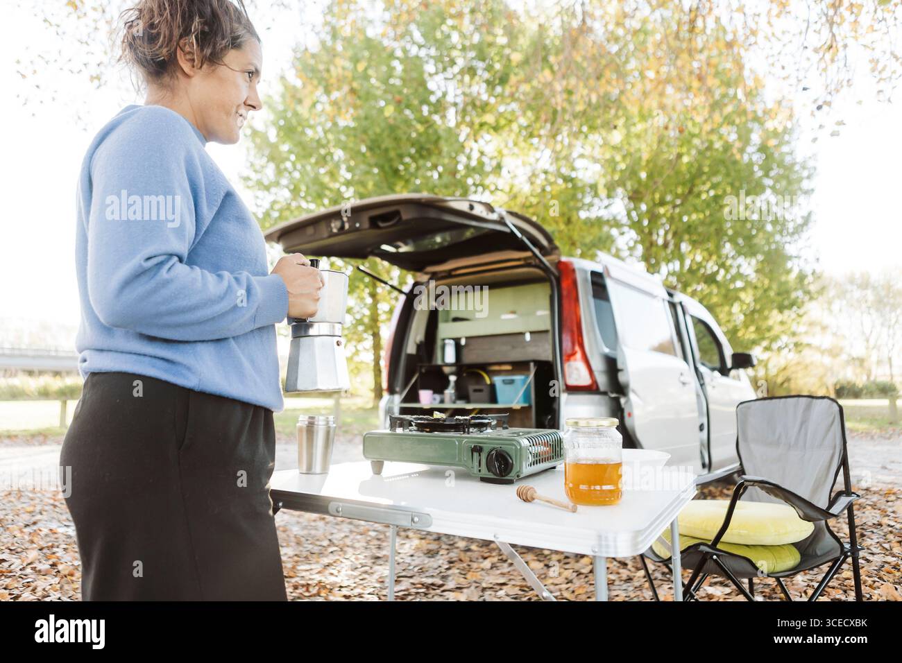 Eine Frau isst morgens ein Frühstück mit Kaffee und Honig neben ihrem Van an einem frischen Herbsttag in Neuseeland. Die Szene fängt das Wesen des Outdoo ein Stockfoto