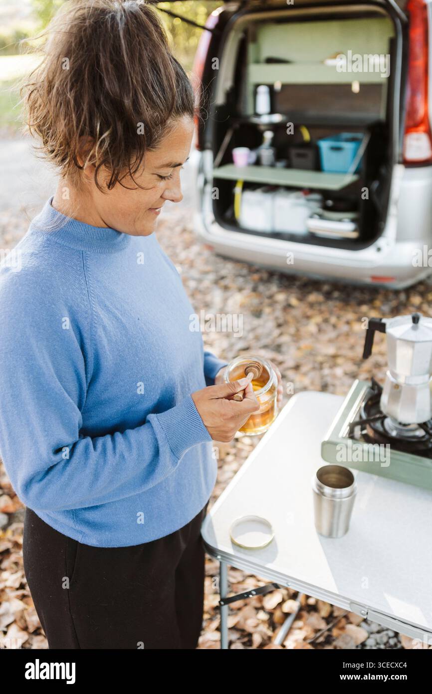 Eine Frau frühstückt im Freien mit ihrem Van in den Herbstwäldern Neuseelands und genießt Kaffee, der mit Honig gesüßt ist. Blätter bedecken den Boden, Crea Stockfoto