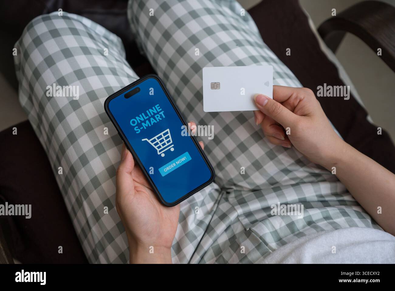Von oben beschnitten nicht erkennbare Person, die ein Smartphone zum Online-Shopping benutzt, und eine Kreditkarte in der anderen Hand hält. Sie sitzt bequem zu Hause Stockfoto