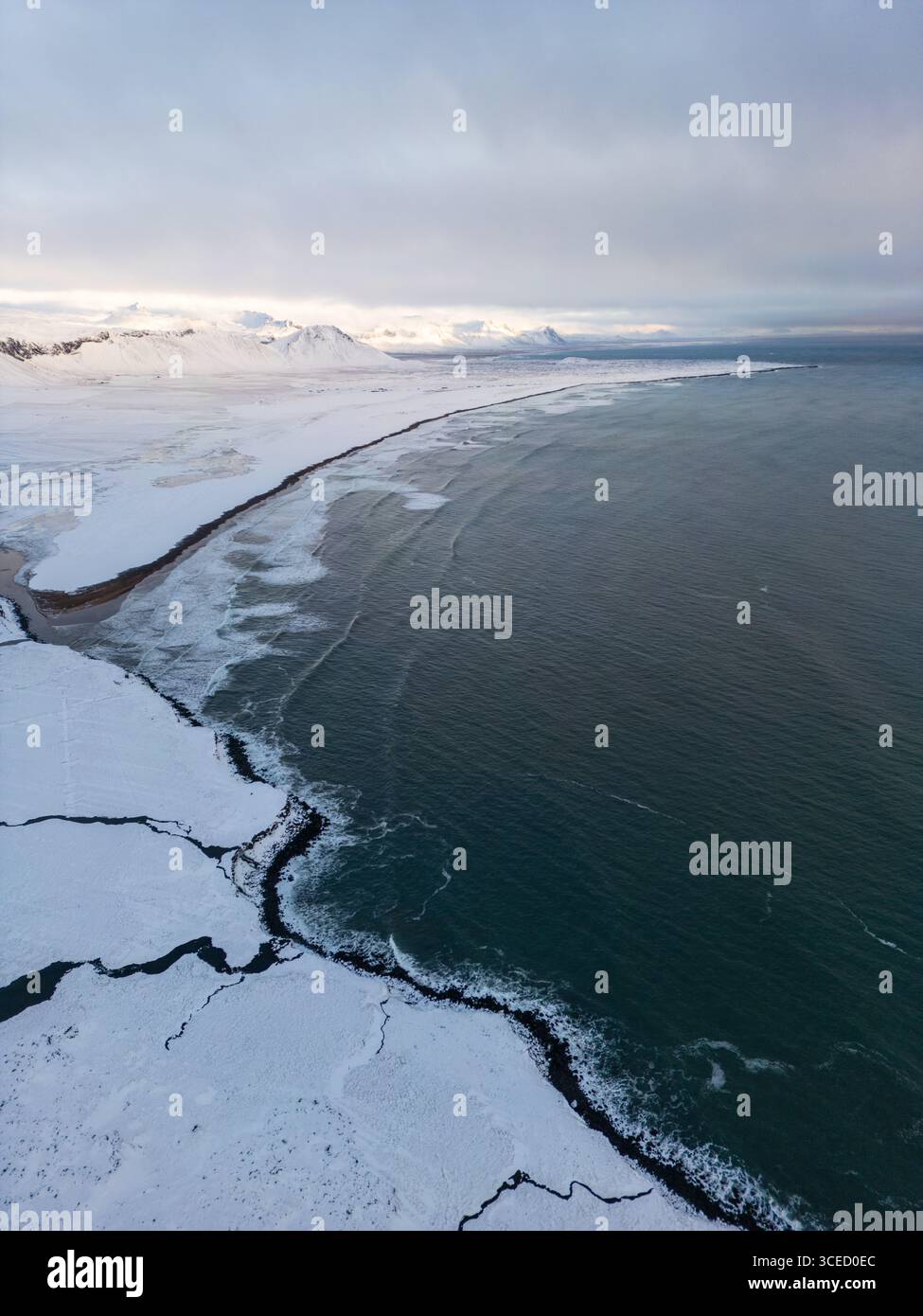 Blick aus der Vogelperspektive auf Islands verschneite Küste, wo zerklüftete Klippen auf den kalten Atlantik treffen. Die weitläufige Winterlandschaft fängt die heitere Schönheit ein Stockfoto
