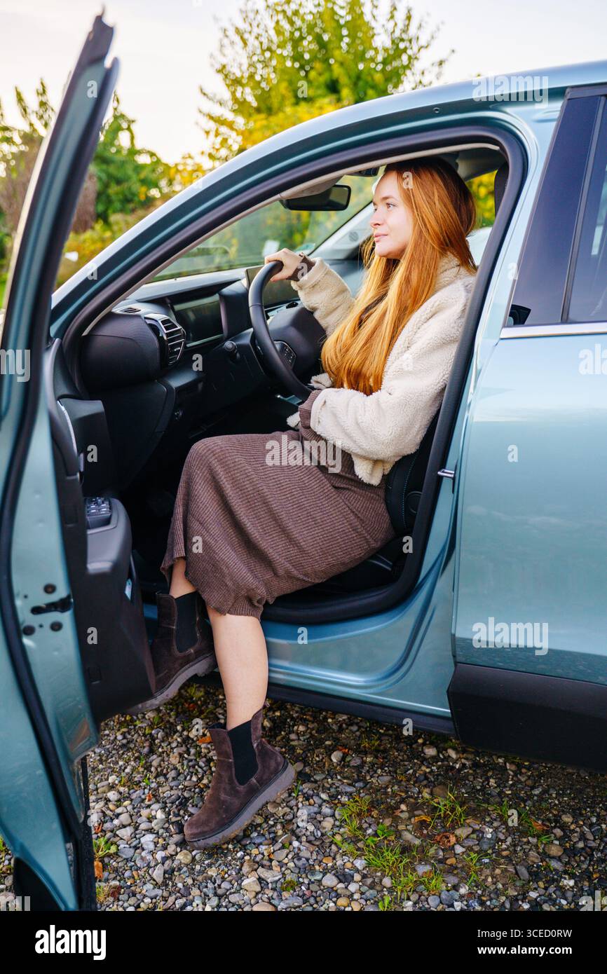 Eine schwangere Frau mit langen roten Haaren sitzt bequem mit geöffneter Tür in einem Auto und genießt die malerische Aussicht draußen. In einem gemütlichen Outfit gekleidet, sie trägt Stockfoto