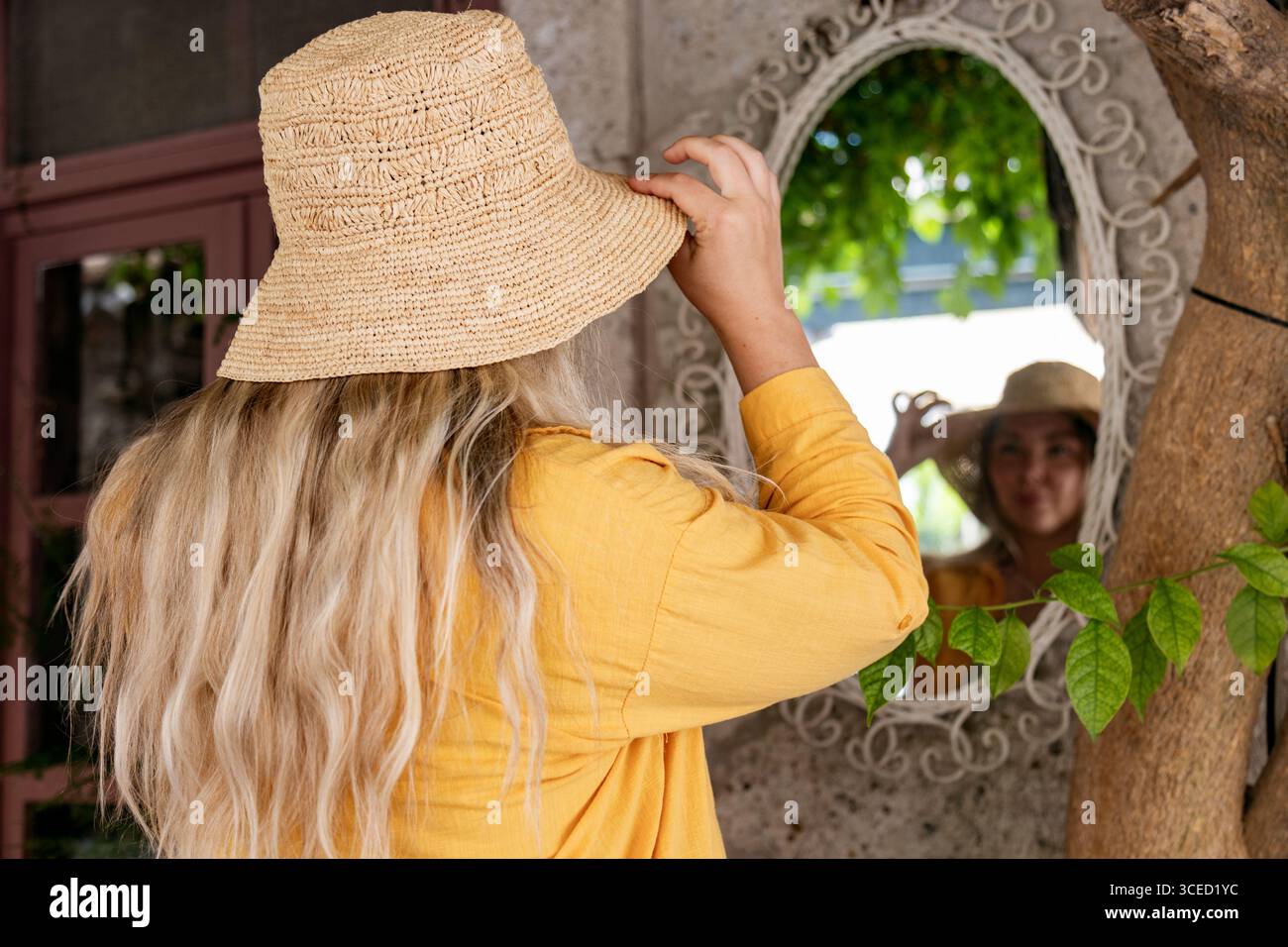 Rückansicht einer jungen Frau in einem gelben Hemd, die ihren Hut vor einem dekorativen Spiegel in einem gemütlichen Café anstellt. Das Ambiente spiegelt ein friedliches Outdo wider Stockfoto
