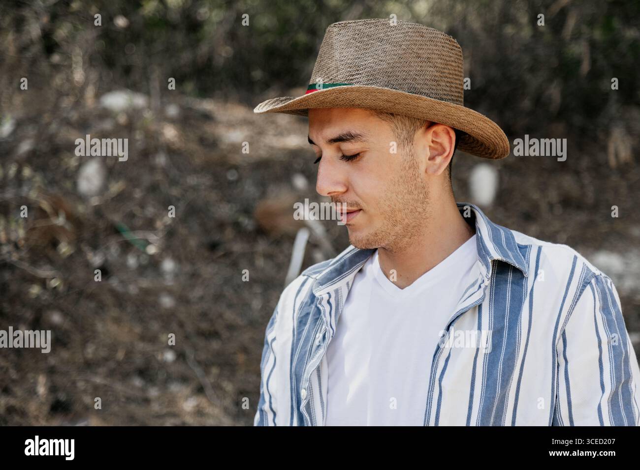 Porträt eines jungen Mannes draußen, der einen Strohhut und ein gestreiftes Hemd trägt. Er steht ruhig vor einem unscharfen natürlichen Hintergrund und fängt einen Moment ein Stockfoto