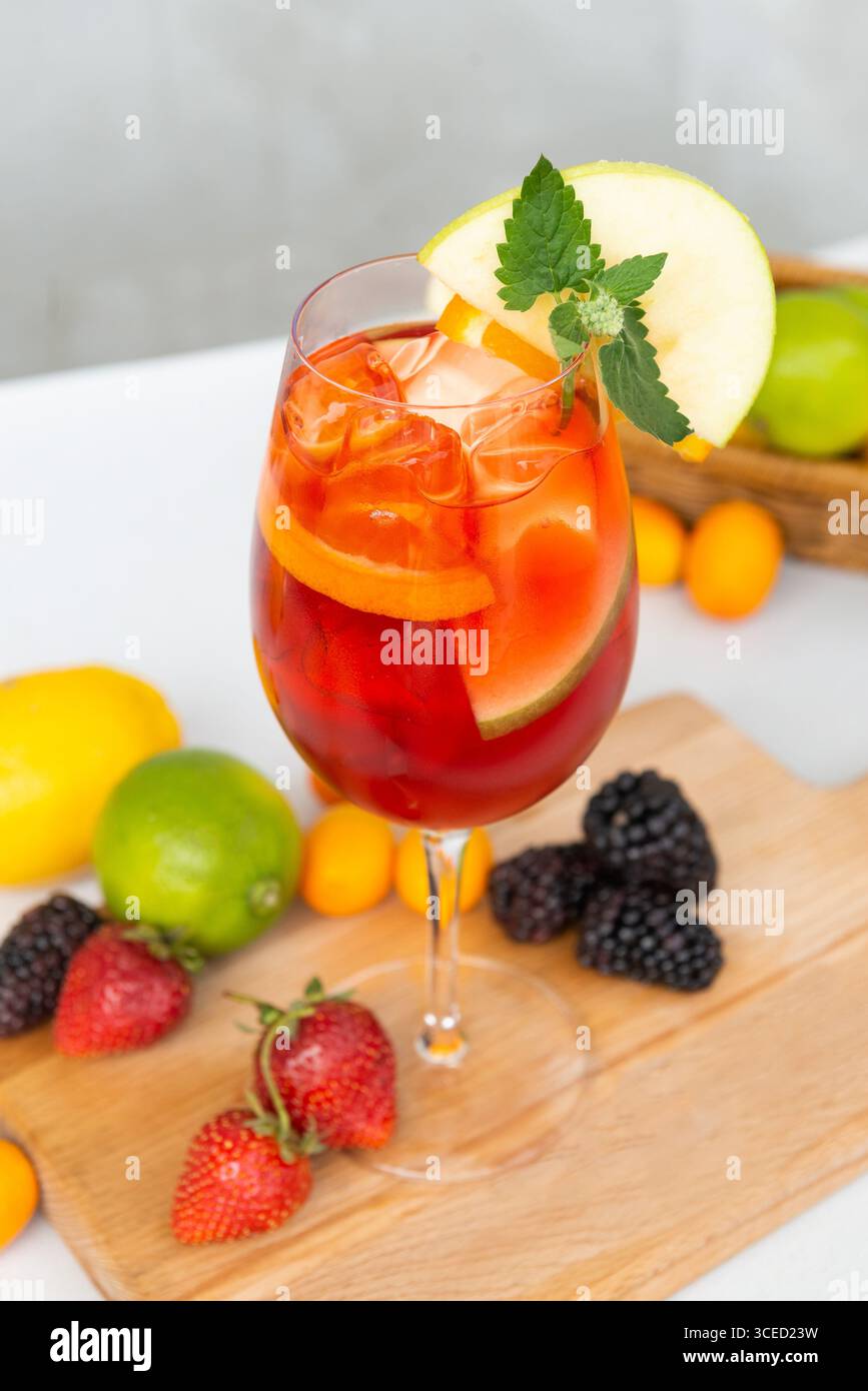 Erfrischendes Sommergetränk in einem Glas mit Eis, garniert mit Apfel und Minze. Umgeben von frischen Früchten wie Erdbeeren, Brombeeren und Zitrusfrüchten Stockfoto