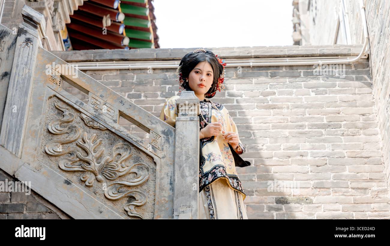 Eine Frau in traditioneller Kleidung der Qing-Dynastie steht auf einer kunstvollen Steintreppe in Pingyao, China, und blickt direkt in die Kamera und reflektiert Stockfoto