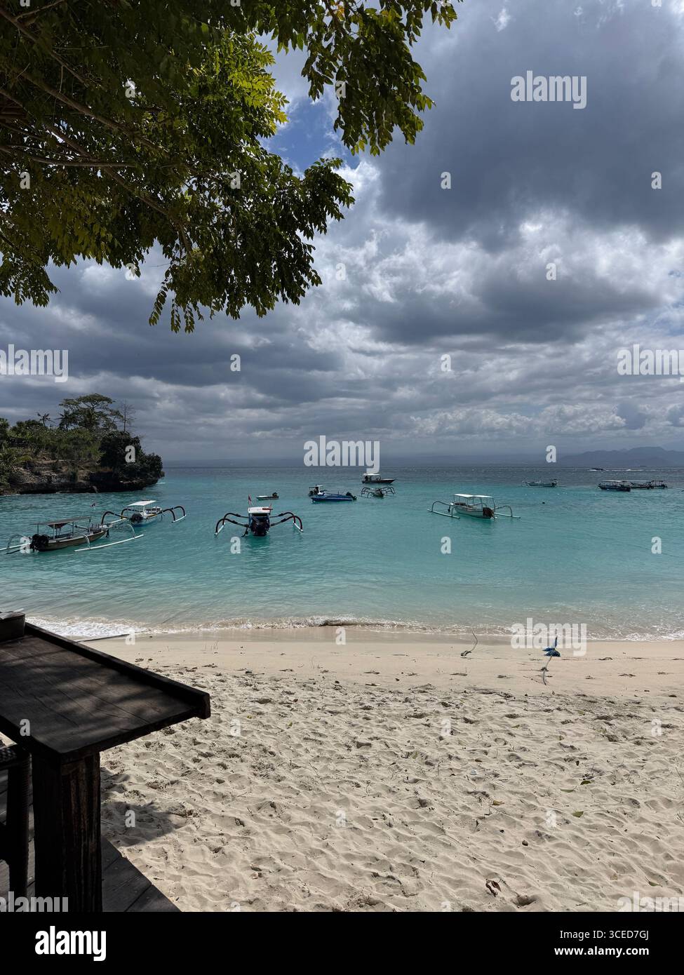 Traditionelle Boote vor Anker auf türkisfarbenem Wasser in der Nähe der Sandküste in Nusa Lembongan, Bali, Indonesien, unter dramatisch bewölktem Himmel und tropischer Atmosphäre - Smartphone-aufgenommenes Stockfoto