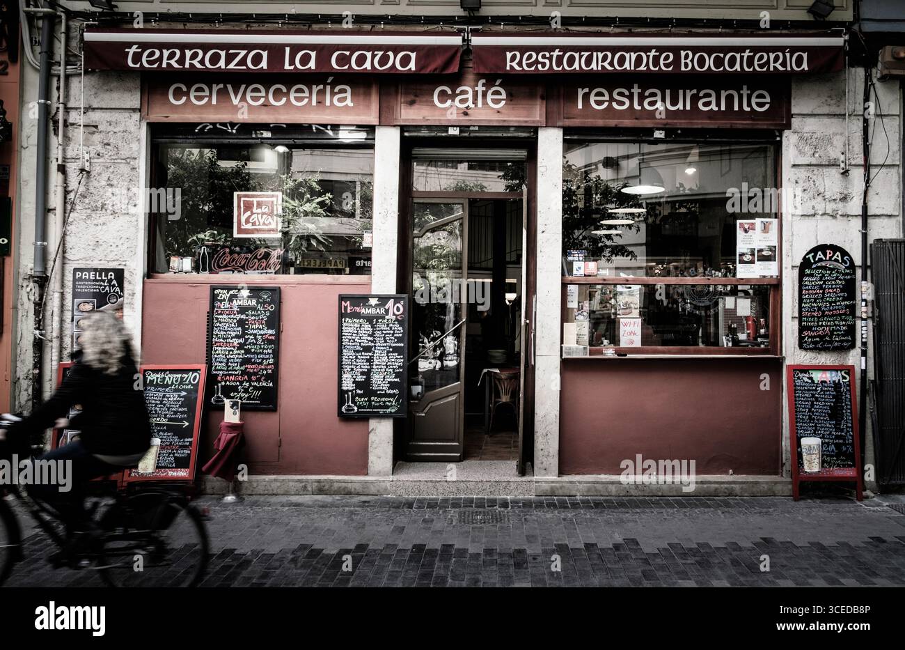 Ein Radfahrer fährt am lebhaften Restaurant und Café Terrazza La Cava vorbei, das Sitzplätze im Freien und Tafelkarten bietet. Das sonnige Ambiente lädt Besucher ein Stockfoto