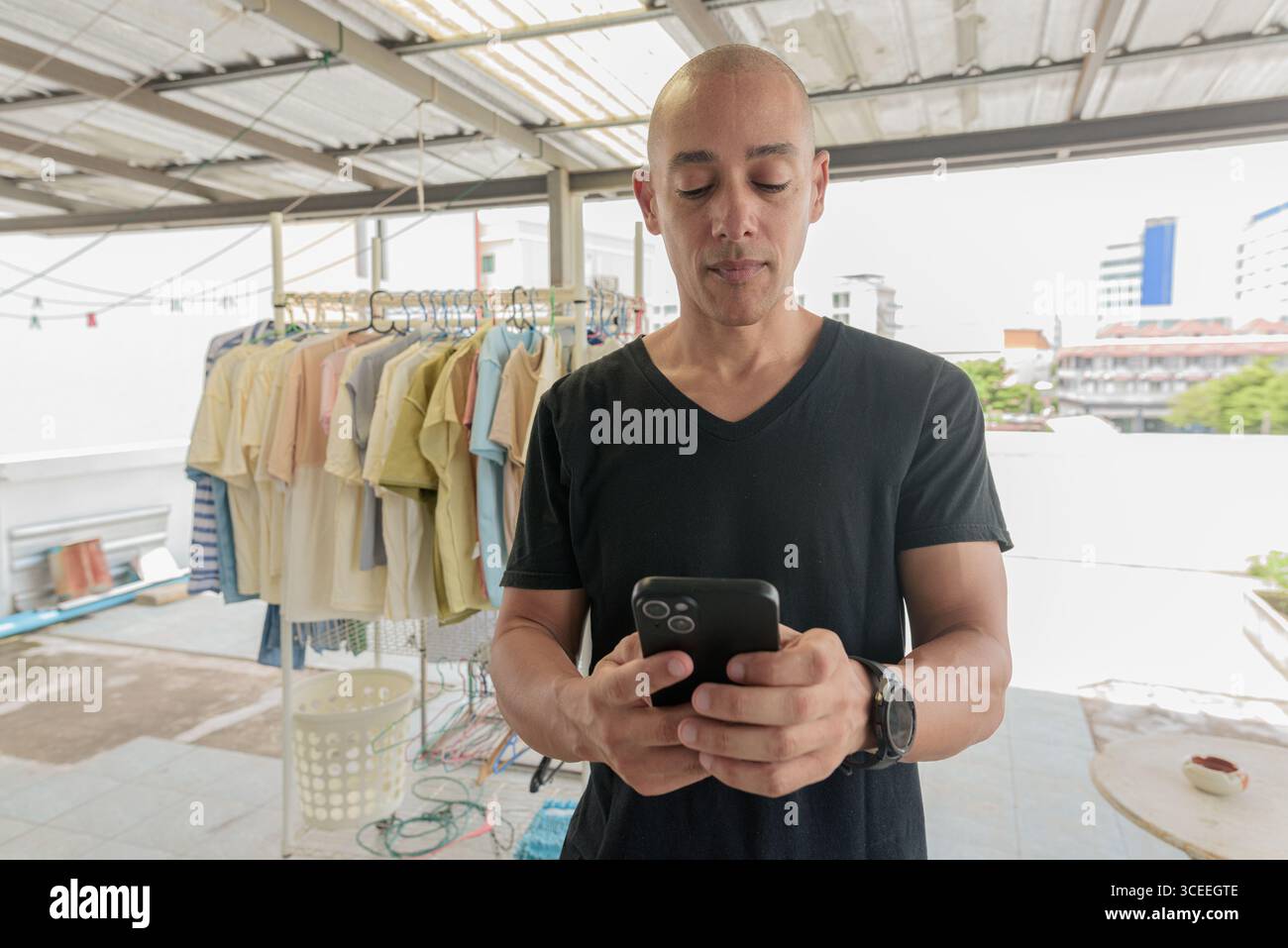 Hispanischer Glatzkopf mit schwarzem T-Shirt, im Freien im Wäschereibereich stehend, Handy beim Waschen gehalten. Multitasking-Konzept, Stockfoto