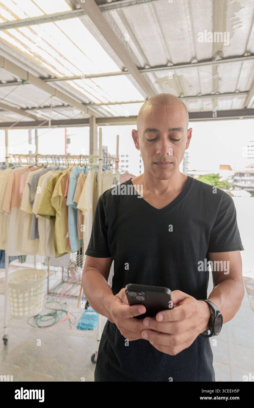 Hispanischer Glatzkopf mit schwarzem T-Shirt, im Freien im Wäschereibereich stehend, Handy beim Waschen gehalten. Multitasking-Konzept, Stockfoto