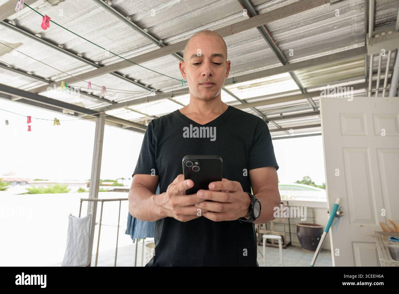 Hispanischer Glatzkopf mit schwarzem T-Shirt, im Freien im Wäschereibereich stehend, Handy beim Waschen gehalten. Multitasking-Konzept, Stockfoto