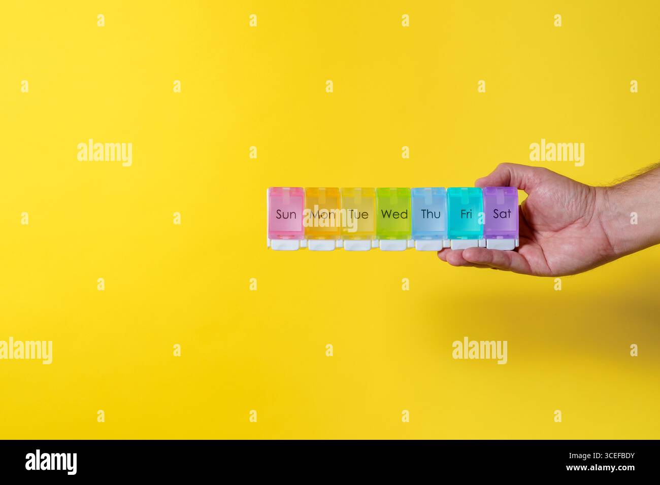 7 Tage wöchentlicher Pille Organizer in Regenbogenfarben mit männlicher Hand auf gelbem Hintergrund mit Kopierraum Stockfoto