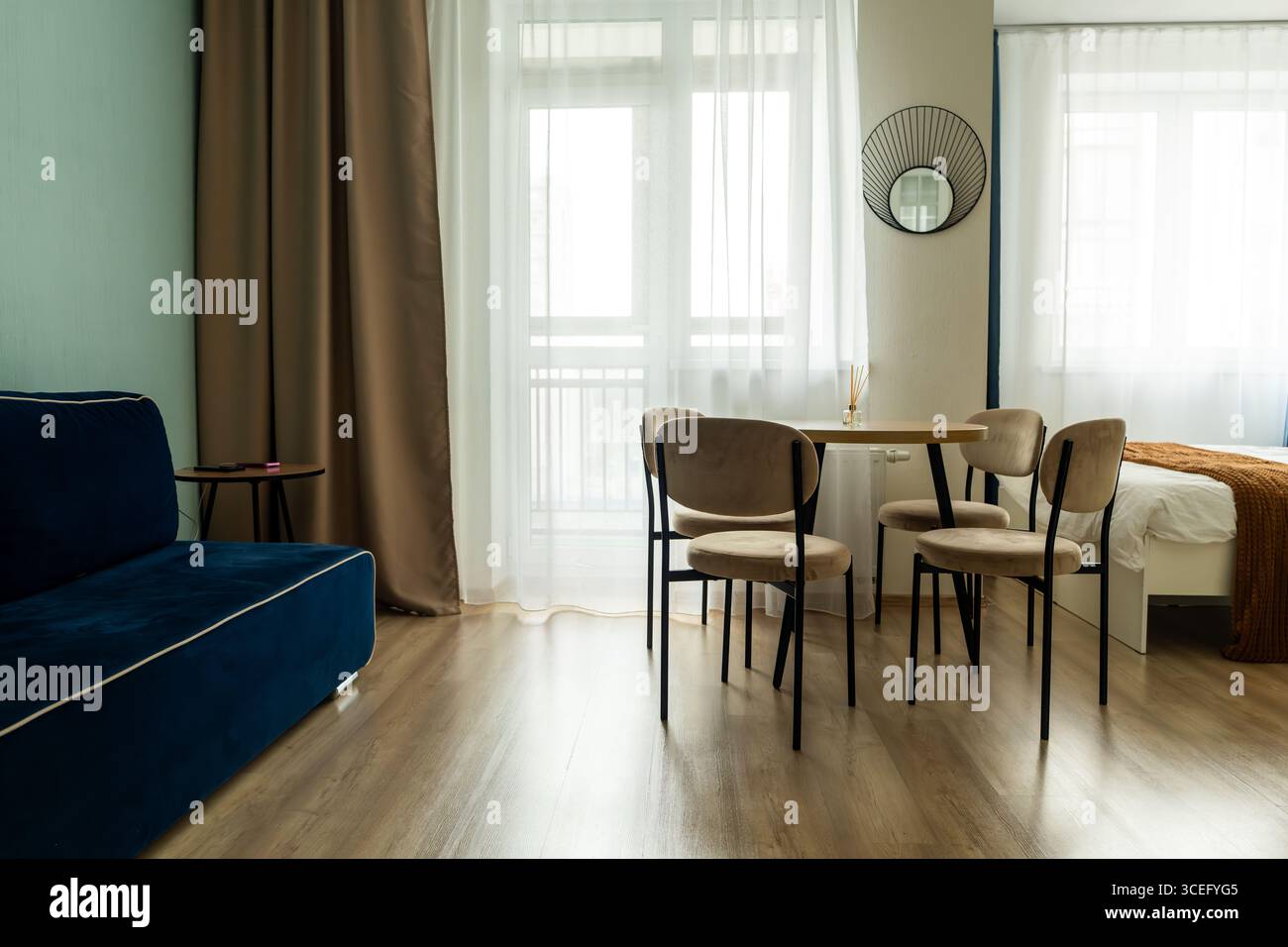 Das Innere eines kleinen Apartments mit Bett, blauen Kissen, hellen Küchenmöbeln, Stühlen mit Tisch, Küchengeräten und Kühlschrank. Apartment Stockfoto