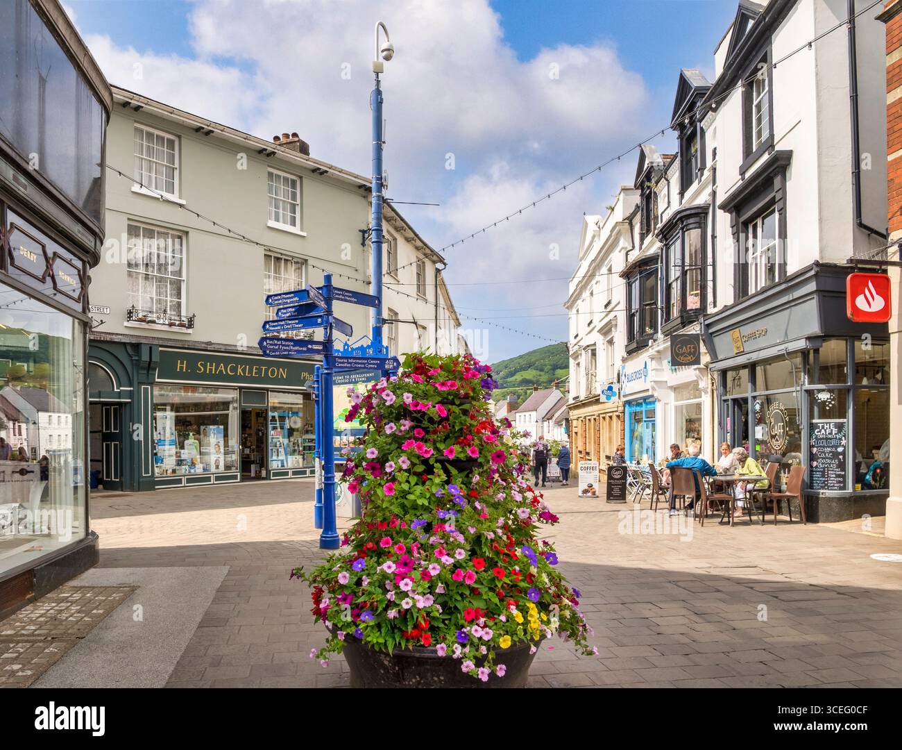 Abergavenny, die High Street, mit Geschäften, Cafés, Menschen, die draußen in der Sonne sitzen, einem Wegweiser für Sehenswürdigkeiten und einem Blumenschmuck. Stockfoto