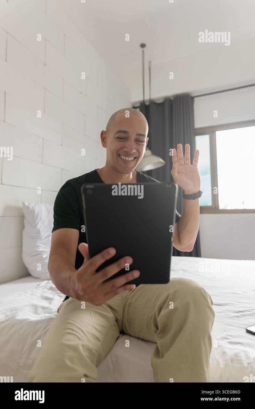 Hispanischer reifer Glatzkopf in den 40er Jahren, der auf dem Bett sitzt, während er einen digitalen Tablet-Computer in seiner modernen Wohnung benutzte, aufgenommen mit Weitwinkelobjektiv Stockfoto