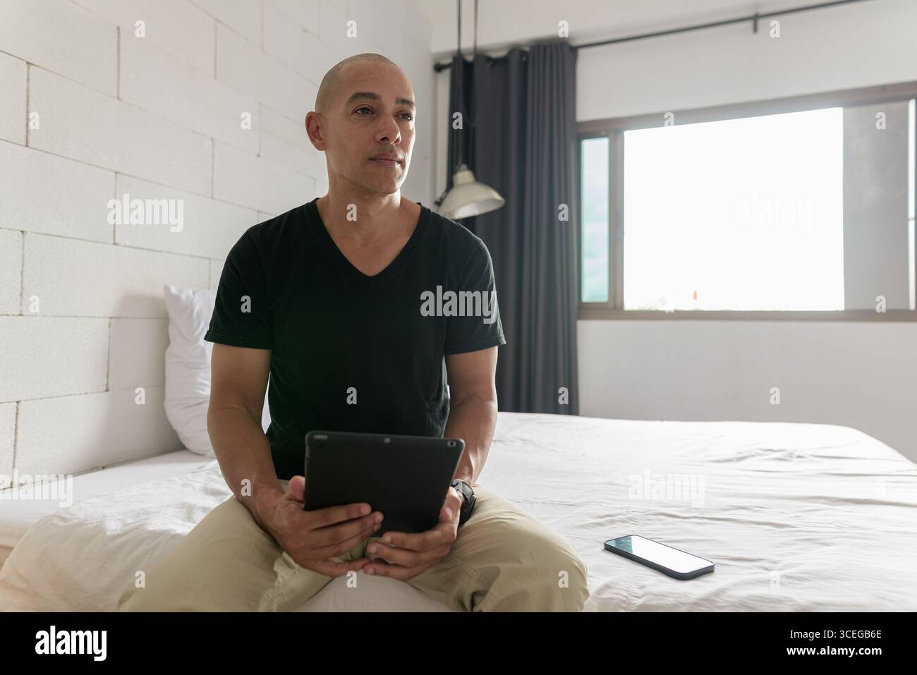 Hispanischer reifer Glatzkopf in den 40er Jahren, der auf dem Bett sitzt, während er einen digitalen Tablet-Computer in seiner modernen Wohnung benutzte, aufgenommen mit Weitwinkelobjektiv Stockfoto