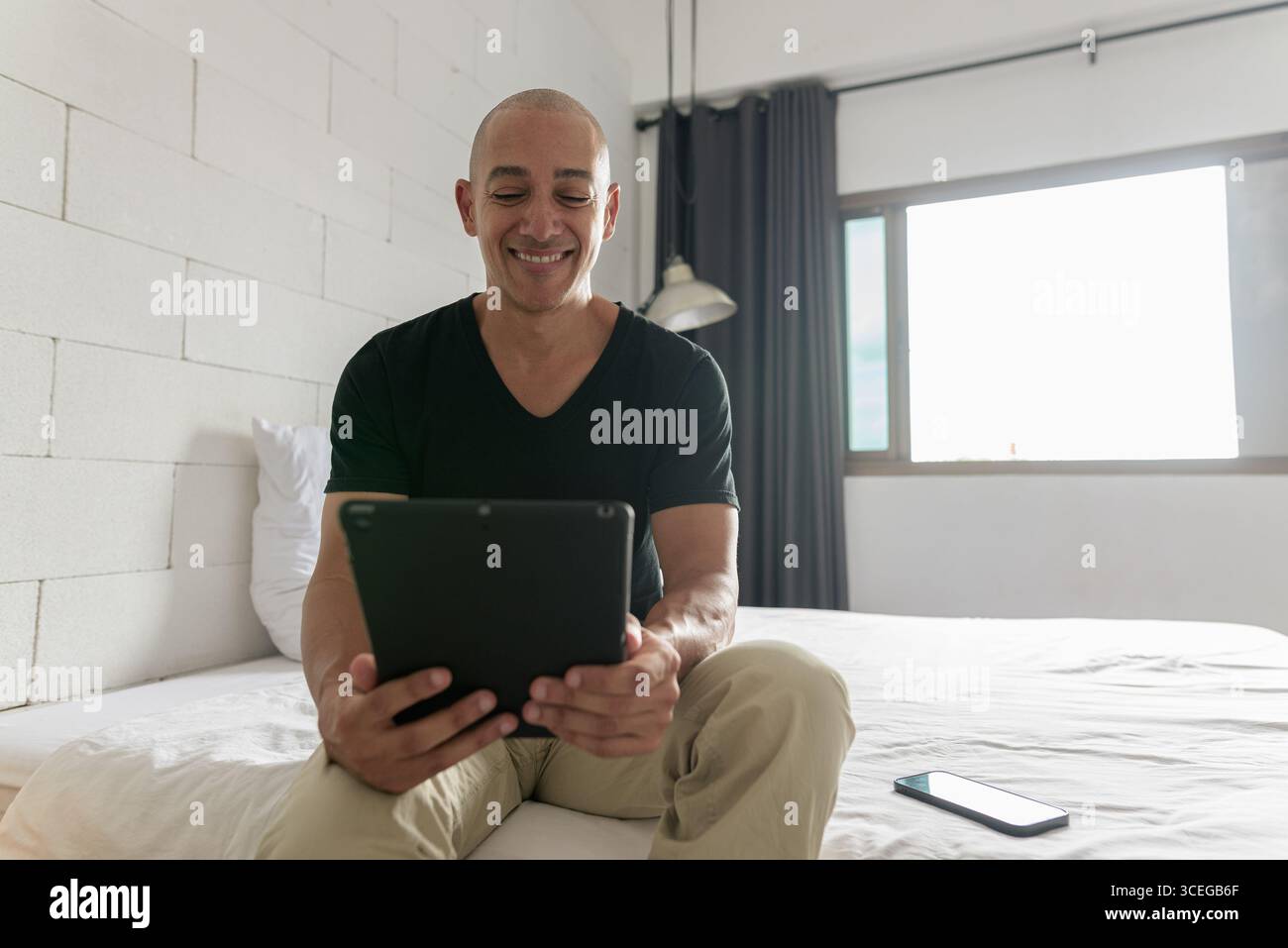 Hispanischer reifer Glatzkopf in den 40er Jahren, der auf dem Bett sitzt, während er einen digitalen Tablet-Computer in seiner modernen Wohnung benutzte, aufgenommen mit Weitwinkelobjektiv Stockfoto