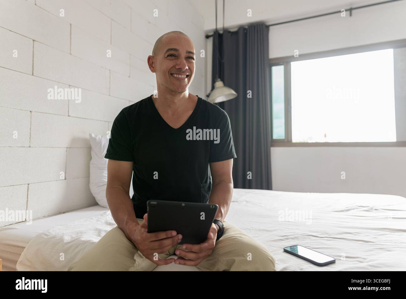 Hispanischer reifer Glatzkopf in den 40er Jahren, der auf dem Bett sitzt, während er einen digitalen Tablet-Computer in seiner modernen Wohnung benutzte, aufgenommen mit Weitwinkelobjektiv Stockfoto