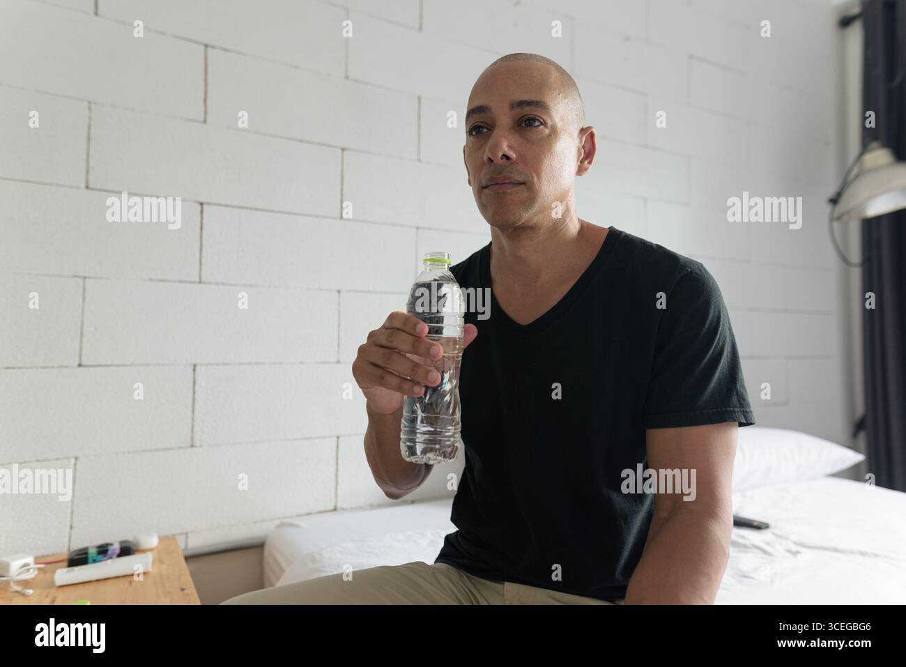 Mittelalter kahler hispanischer Mann in schwarzem T-Shirt, der Wasser aus einer Plastikflasche in einem minimalistischen Apartment trinkt. Hydratation in Innenräumen und gesundes lifestyl Stockfoto