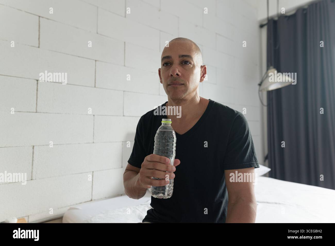 Mittelalter kahler hispanischer Mann in schwarzem T-Shirt, der Wasser aus einer Plastikflasche in einem minimalistischen Apartment trinkt. Hydratation in Innenräumen und gesundes lifestyl Stockfoto