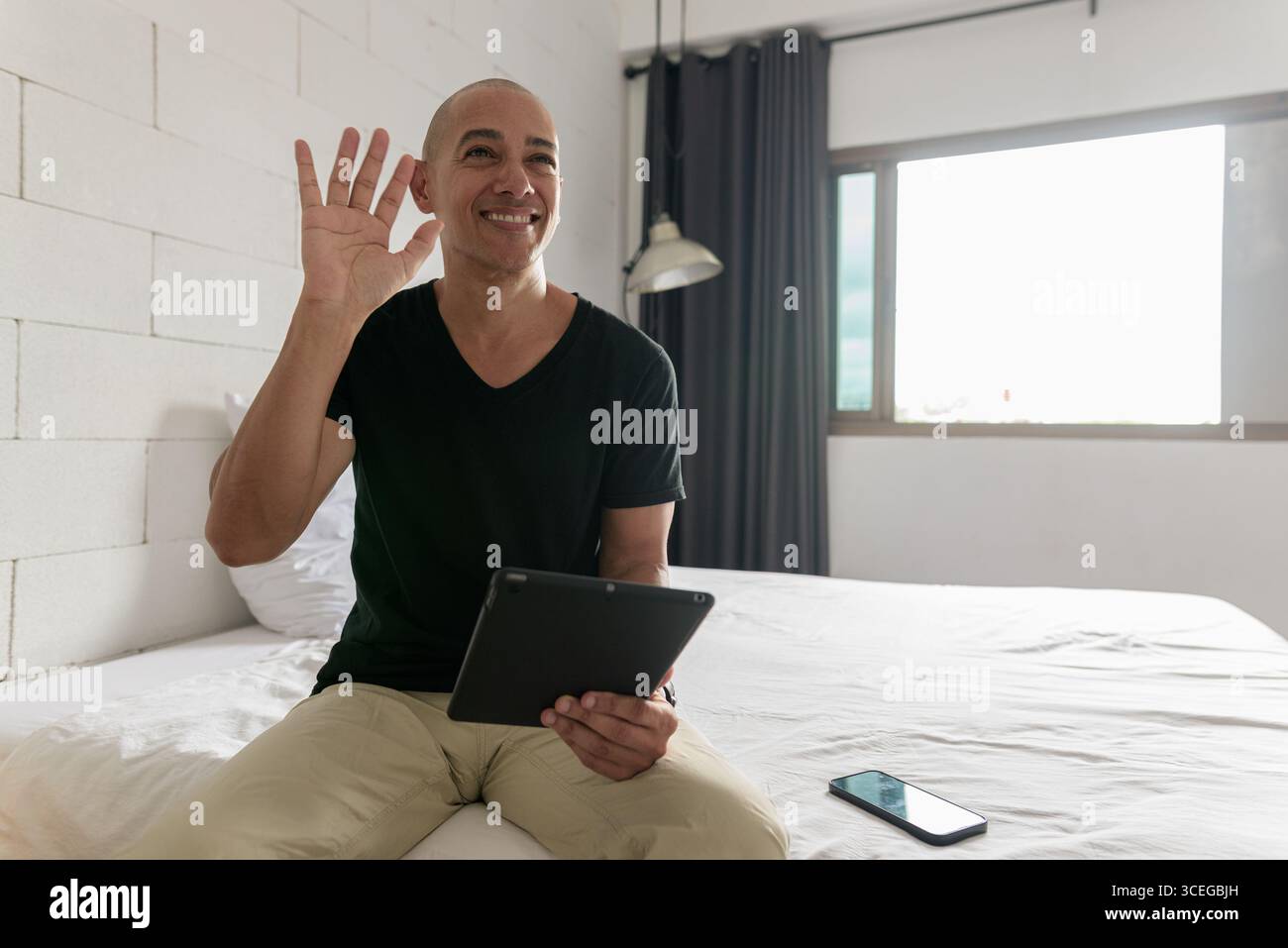 Hispanischer reifer Glatzkopf in den 40er Jahren, der auf dem Bett sitzt, während er einen digitalen Tablet-Computer in seiner modernen Wohnung benutzte, aufgenommen mit Weitwinkelobjektiv Stockfoto