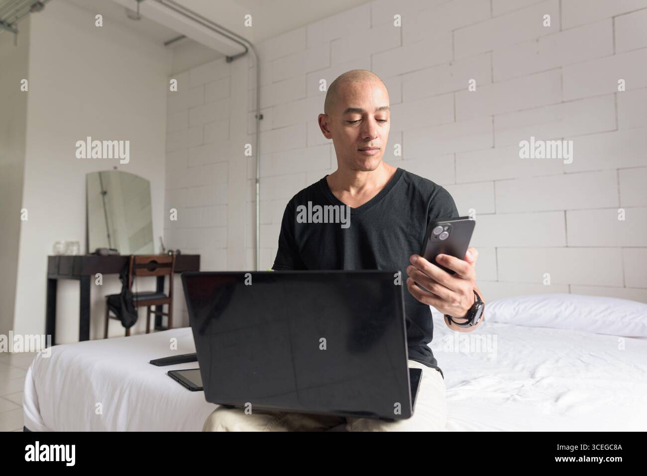 Ein Mann mit einem glatten hispanischen Mann mittleren Alters, der ein schwarzes T-Shirt trägt und auf dem Bett sitzt, während er einen Laptop und ein Telefon in einem minimalistischen weißen Apartment benutzte. Modernes Zuhause Stockfoto
