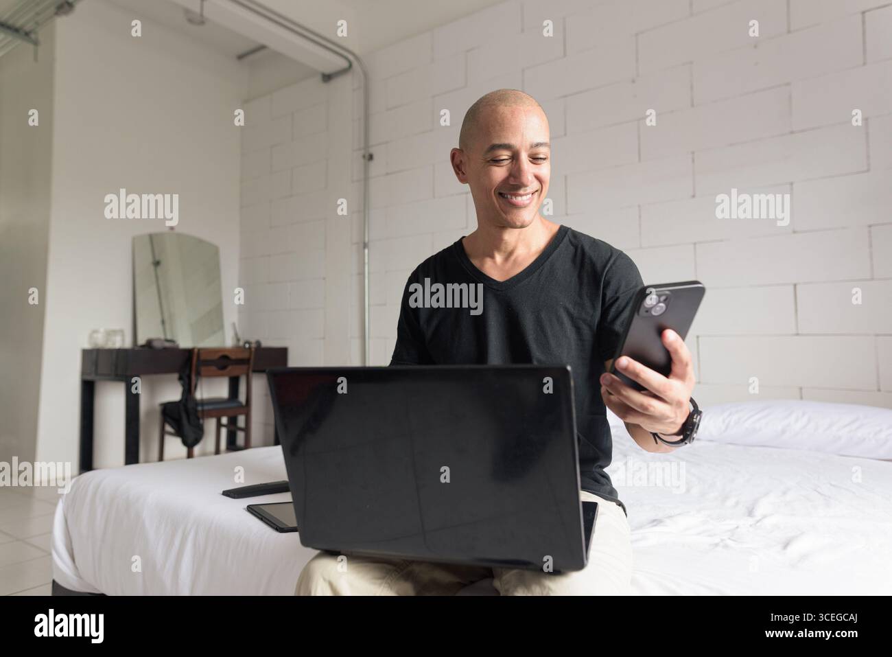 Ein Mann mit einem glatten hispanischen Mann mittleren Alters, der ein schwarzes T-Shirt trägt und auf dem Bett sitzt, während er einen Laptop und ein Telefon in einem minimalistischen weißen Apartment benutzte. Modernes Zuhause Stockfoto
