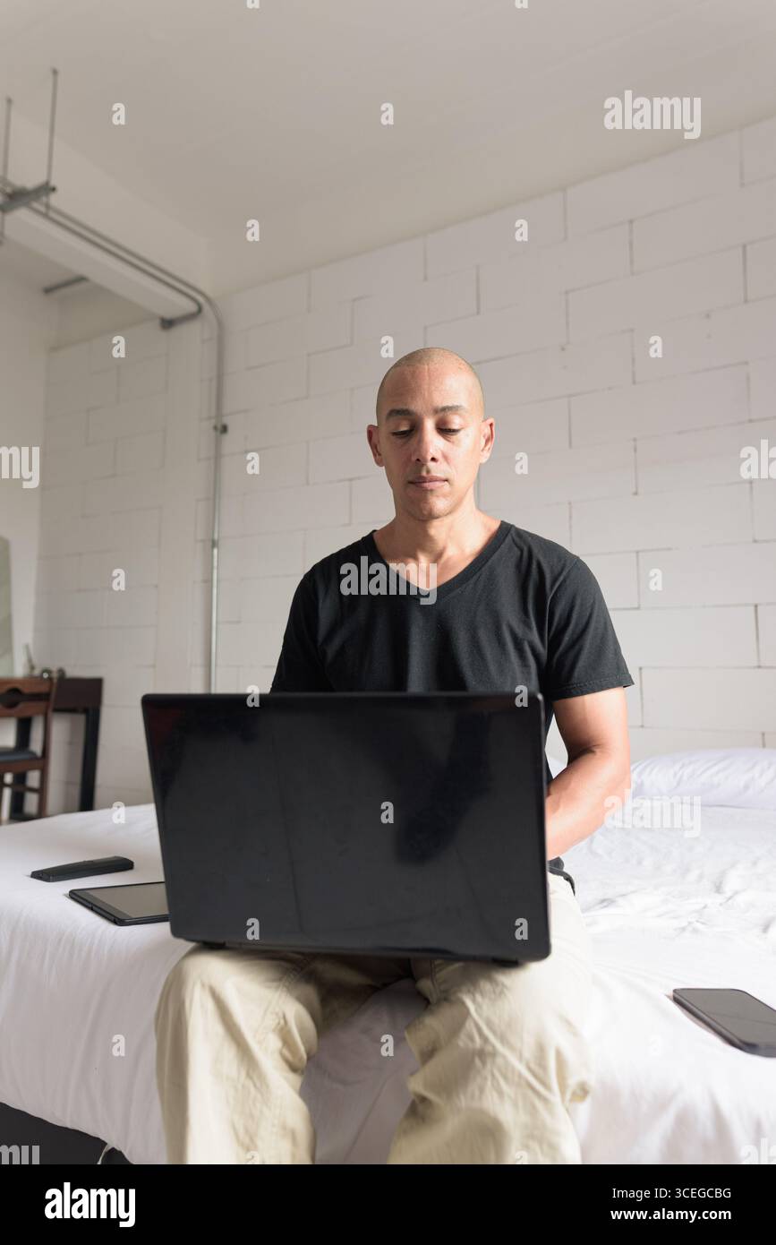 Ein Mann aus dem mittelalten, glatten Hispanic mit schwarzem T-Shirt, der auf dem Bett sitzt und einen Laptop in einem minimalistischen weißen Apartment benutzt. Modernes Home Working li Stockfoto