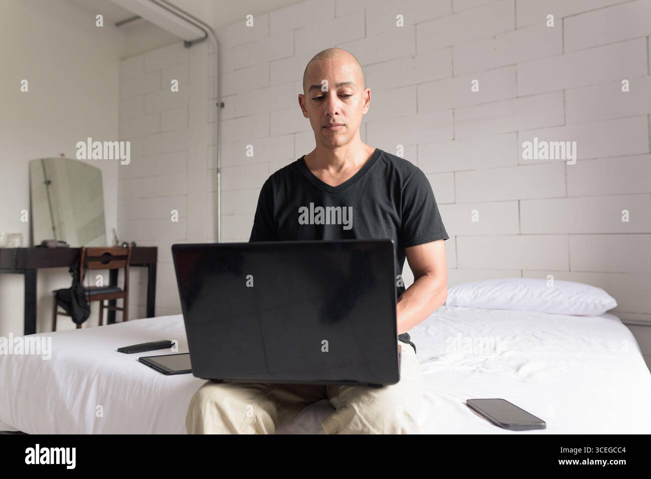 Ein Mann aus dem mittelalten, glatten Hispanic mit schwarzem T-Shirt, der auf dem Bett sitzt und einen Laptop in einem minimalistischen weißen Apartment benutzt. Modernes Home Working li Stockfoto