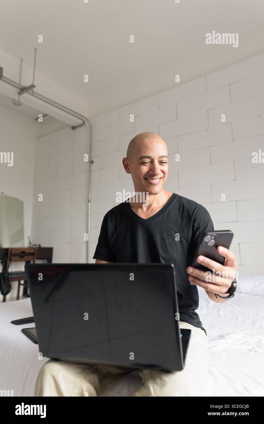Ein Mann mit einem glatten hispanischen Mann mittleren Alters, der ein schwarzes T-Shirt trägt und auf dem Bett sitzt, während er einen Laptop und ein Telefon in einem minimalistischen weißen Apartment benutzte. Modernes Zuhause Stockfoto