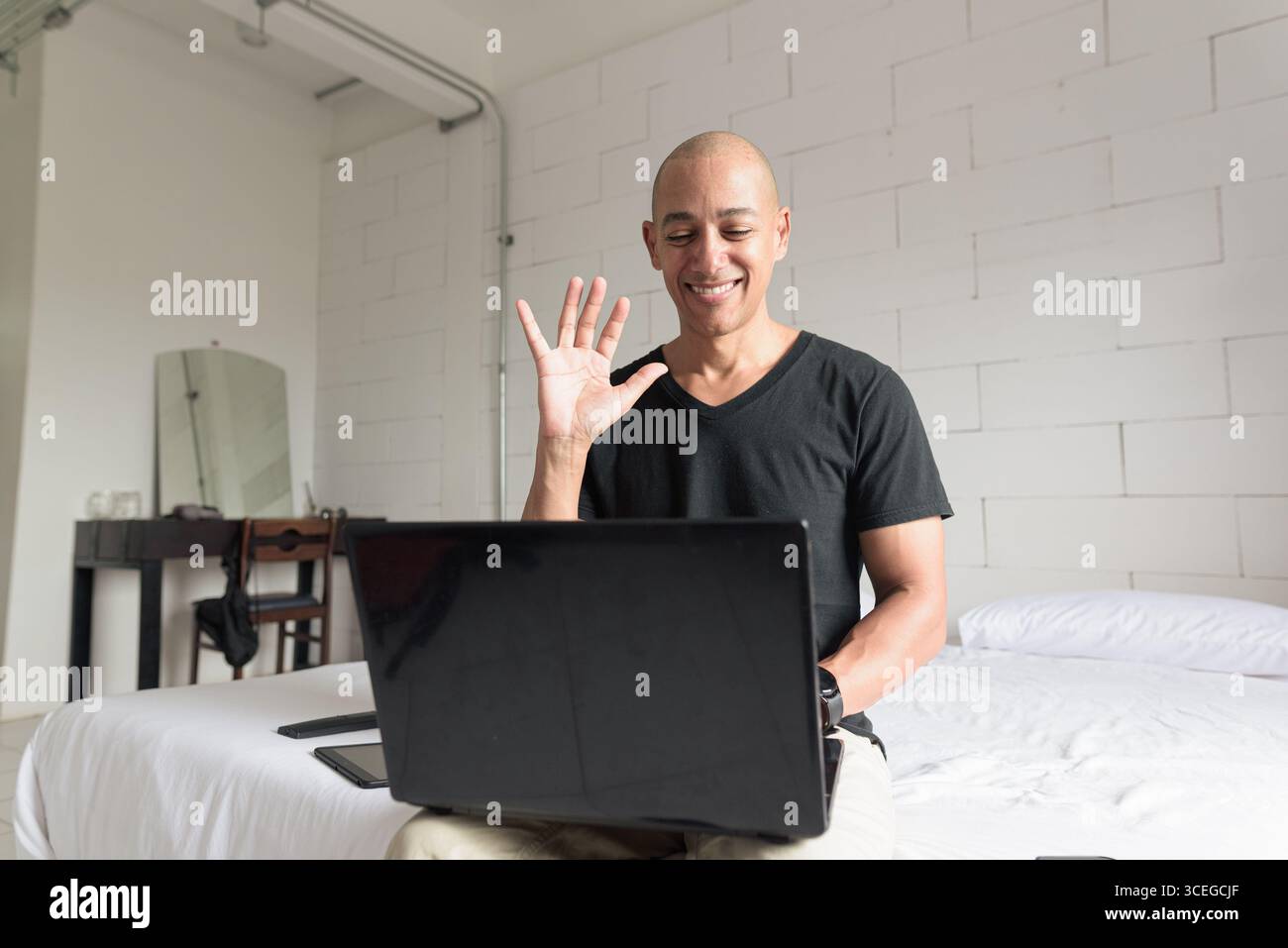 Ein Mann aus dem mittelalten, glatten Hispanic mit schwarzem T-Shirt, der auf dem Bett sitzt und einen Laptop in einem minimalistischen weißen Apartment benutzt. Modernes Home Working li Stockfoto