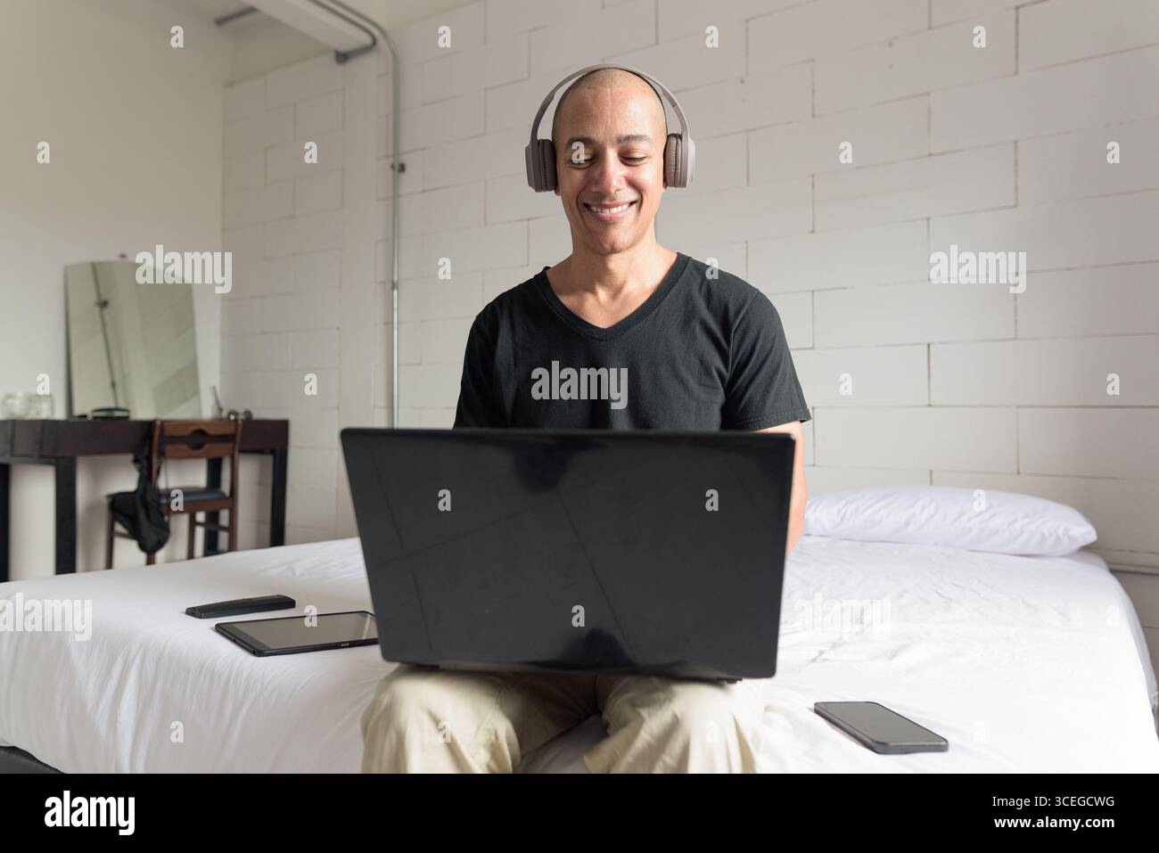 Ein Mann aus dem mittelalten, glatten Hispanic mit schwarzem T-Shirt, der auf dem Bett sitzt und einen Laptop in einem minimalistischen weißen Apartment benutzt. Modernes Home Working li Stockfoto