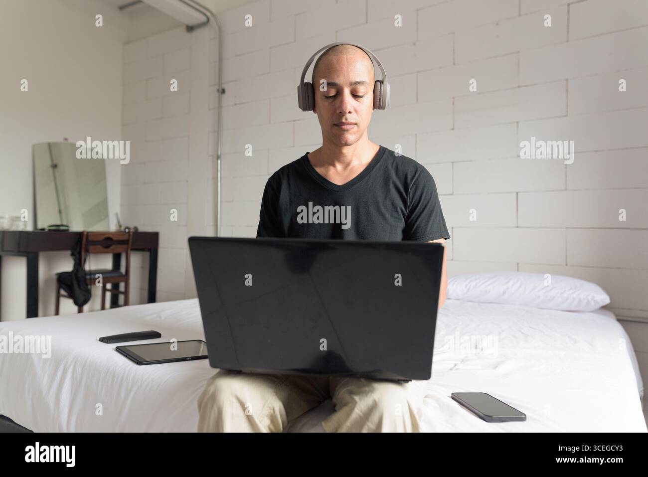 Ein Mann aus dem mittelalten, glatten Hispanic mit schwarzem T-Shirt, der auf dem Bett sitzt und einen Laptop in einem minimalistischen weißen Apartment benutzt. Modernes Home Working li Stockfoto