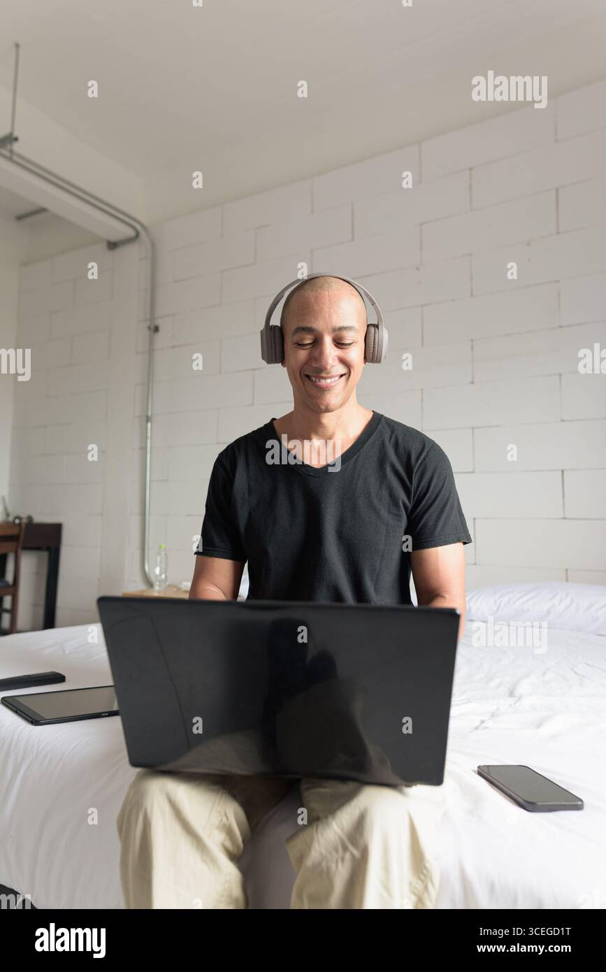 Ein Mann aus dem mittelalten, glatten Hispanic mit schwarzem T-Shirt, der auf dem Bett sitzt und einen Laptop in einem minimalistischen weißen Apartment benutzt. Modernes Home Working li Stockfoto
