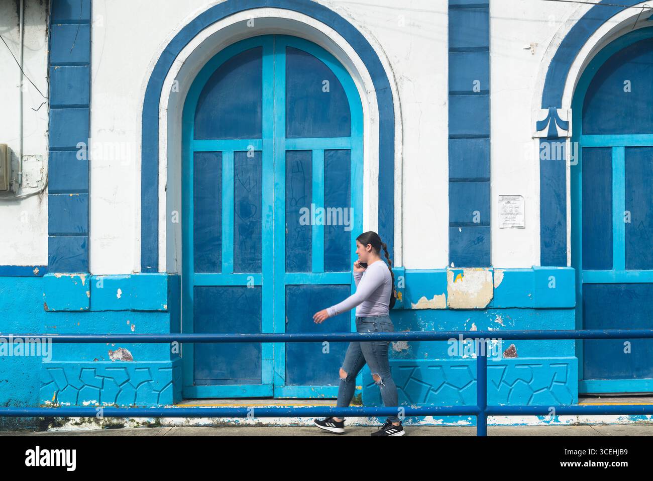 Junge Dame mit Handy, die an einem blau-weiß bemalten Kolonialhaus in Filandia, Departement Quindío, Kolumbien vorbeiläuft Stockfoto
