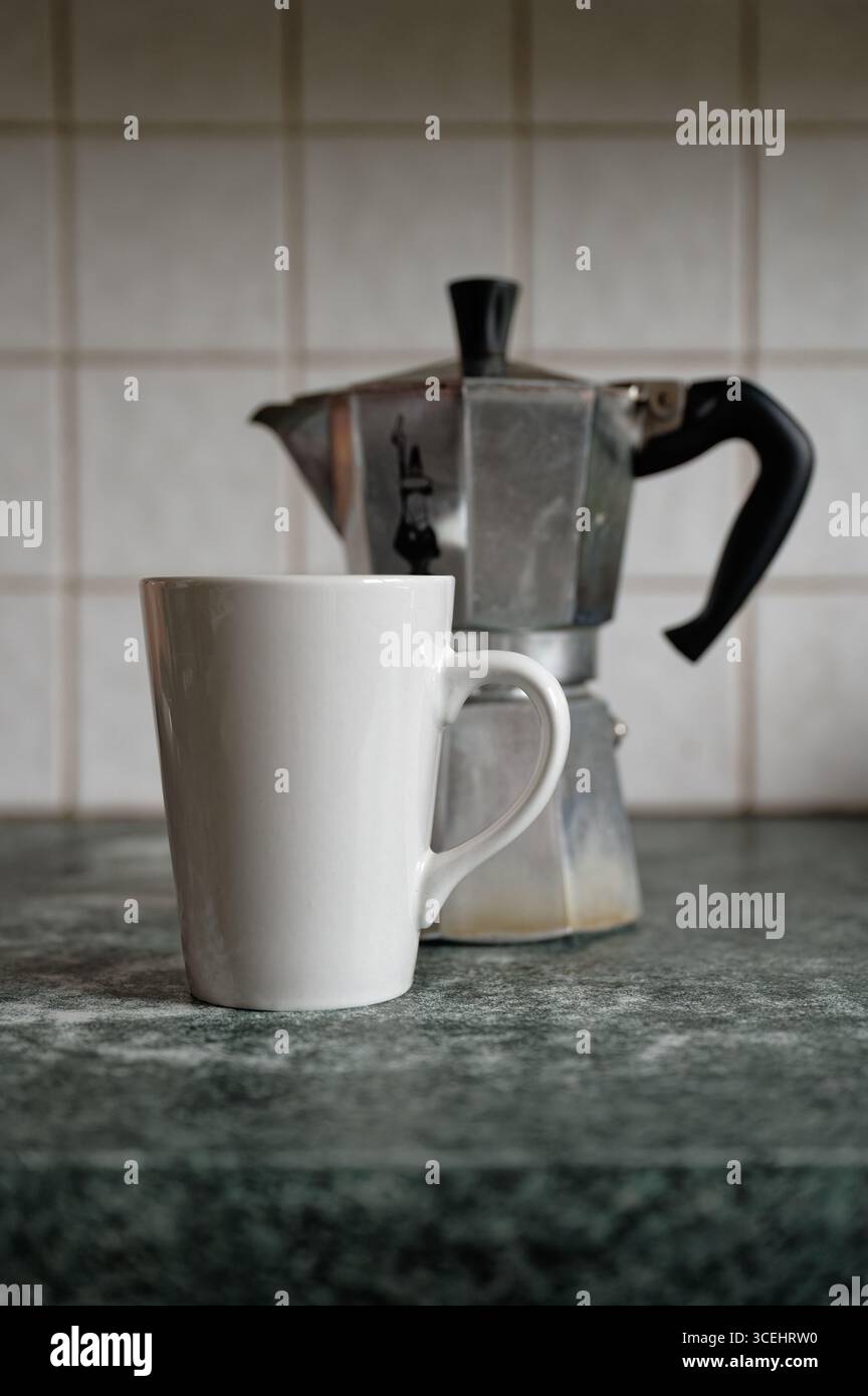 Einfarbige weiße Tasse vor einer kleinen Bialetti Moka Express Kaffeemaschine auf einer grün-grau gemusterten Küchenzeile mit weißen Fliesen im Hintergrund. Stockfoto