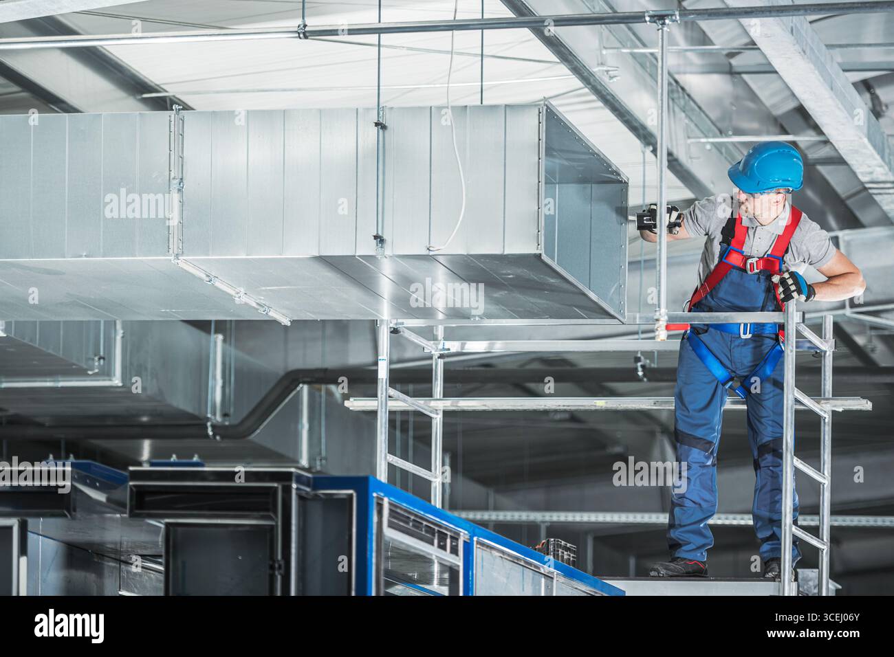 Ein erfahrener HLK-Mitarbeiter balanciert auf einem Gerüst aus und sichert einen Luftkanal in einem großen Lager sorgfältig. Helle Deckenleuchten beleuchten den Raum. Stockfoto
