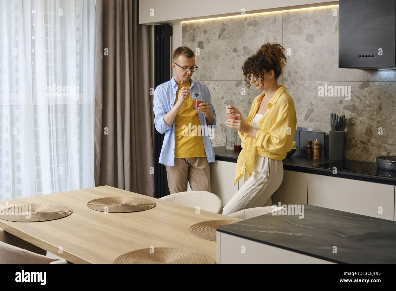 Ein Paar isst einen Frucht-Smoothie, während er in der Küche steht. Der stilvolle Raum verfügt über einen hölzernen Esstisch, moderne Geräte und sanfte Beleuchtung, Stockfoto