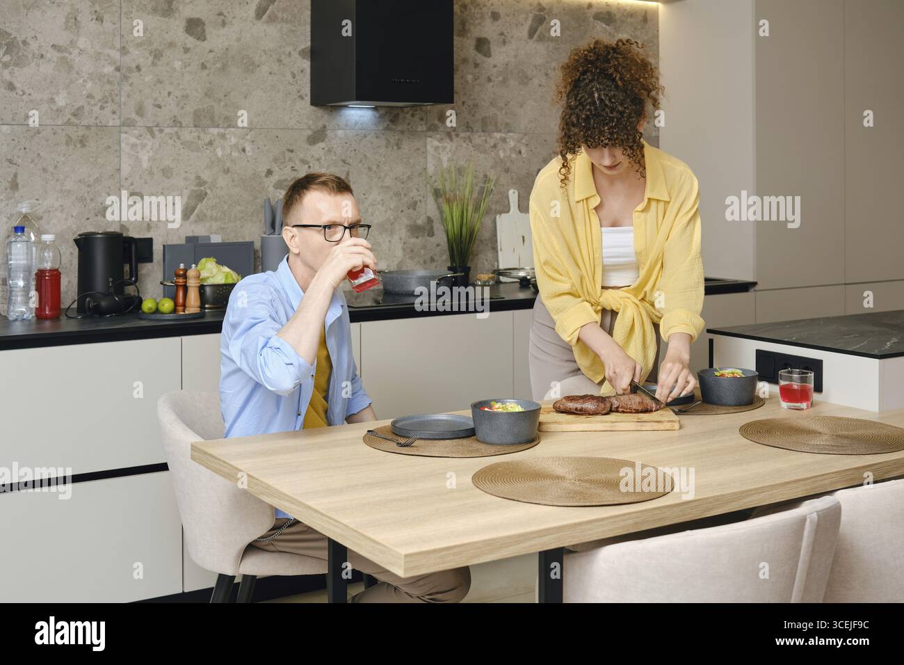 Ein Mann sitzt an einem hölzernen Esstisch und schlürft einen Drink, während eine Frau auf der Arbeitsplatte in einer stilvollen Küche eine Mahlzeit zubereitet. Die Atmosphäre ist gemütlich und Stockfoto
