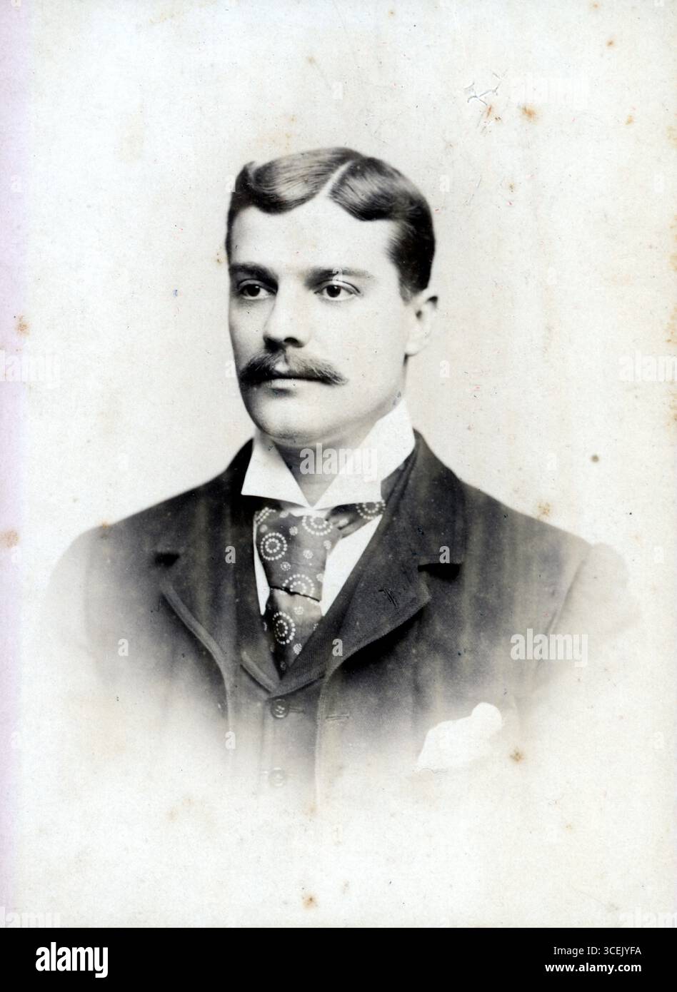 Ein gepflegter Herr posiert in formeller Kleidung – dunkle Jacke, Weste und knuspriges weißes Hemd – mit gemusterter Krawatte und einem ordentlich gefalteten Taschenfeld. Seine geteilten Haare und sein raffinierter Schnurrbart spiegeln die Pflegestandards des späten 19. Bis frühen 20. Jahrhunderts wider. Die verblassten Kanten und die leichten Flecken verleihen dem Foto Archivstruktur, während der schlichte Hintergrund die Aufmerksamkeit auf seinen komponierten Ausdruck lenkt. Porträt vermittelt stille Selbstvertrauen und schicksalhafte Eleganz, symbolisch für die zeitweiligen Edwardianischen Studiokonventionen. Aus einer Reihe von originalen Albumenfotos aus dem späten viktorianischen und frühen Edwardianischen Stil. Stockfoto