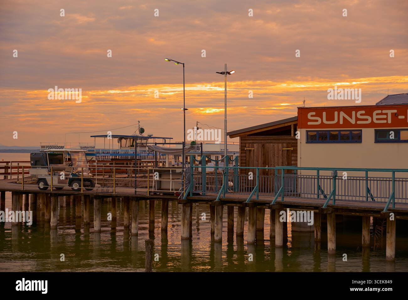 Warmer Sonnenuntergang über dem Pier-Café mit dem „Sunset“-Schild, leuchtenden Lampen und einem Golfwagen, der im goldenen Licht geparkt ist. Stockfoto
