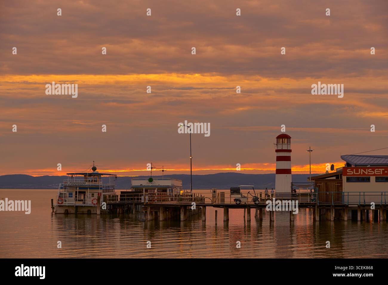 Sunset Bar Pier mit Golfwagen und Anlegeboot unter orangegrauem Himmel. Die Lampen leuchten warm, reflektieren auf dem See, mit einem Leuchtturm am Horizont. Stockfoto