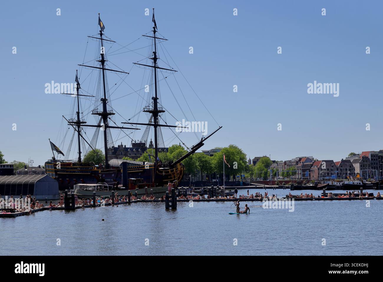 Ein Outdoor-Schwimmbereich am Marineterrein mit Menschenmassen auf schwimmenden Plattformen und einem historischen Großschiff, das in der Nähe von Amsterdam, Niederlande, bei Tag vor Anker liegt. Stockfoto