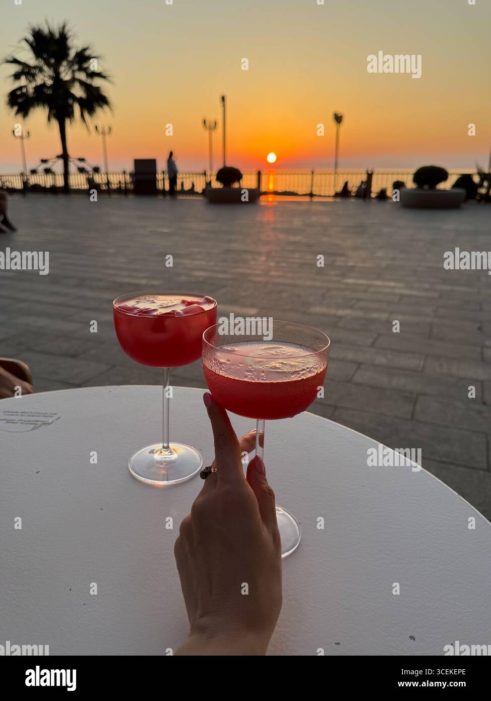 Das Bild zeigt zwei rote Cocktails in Stielgläsern auf einem weißen runden Tisch. Eine der Gläser wird von einer Hand mit bemalten Nägeln und einem Ring gehalten Stockfoto