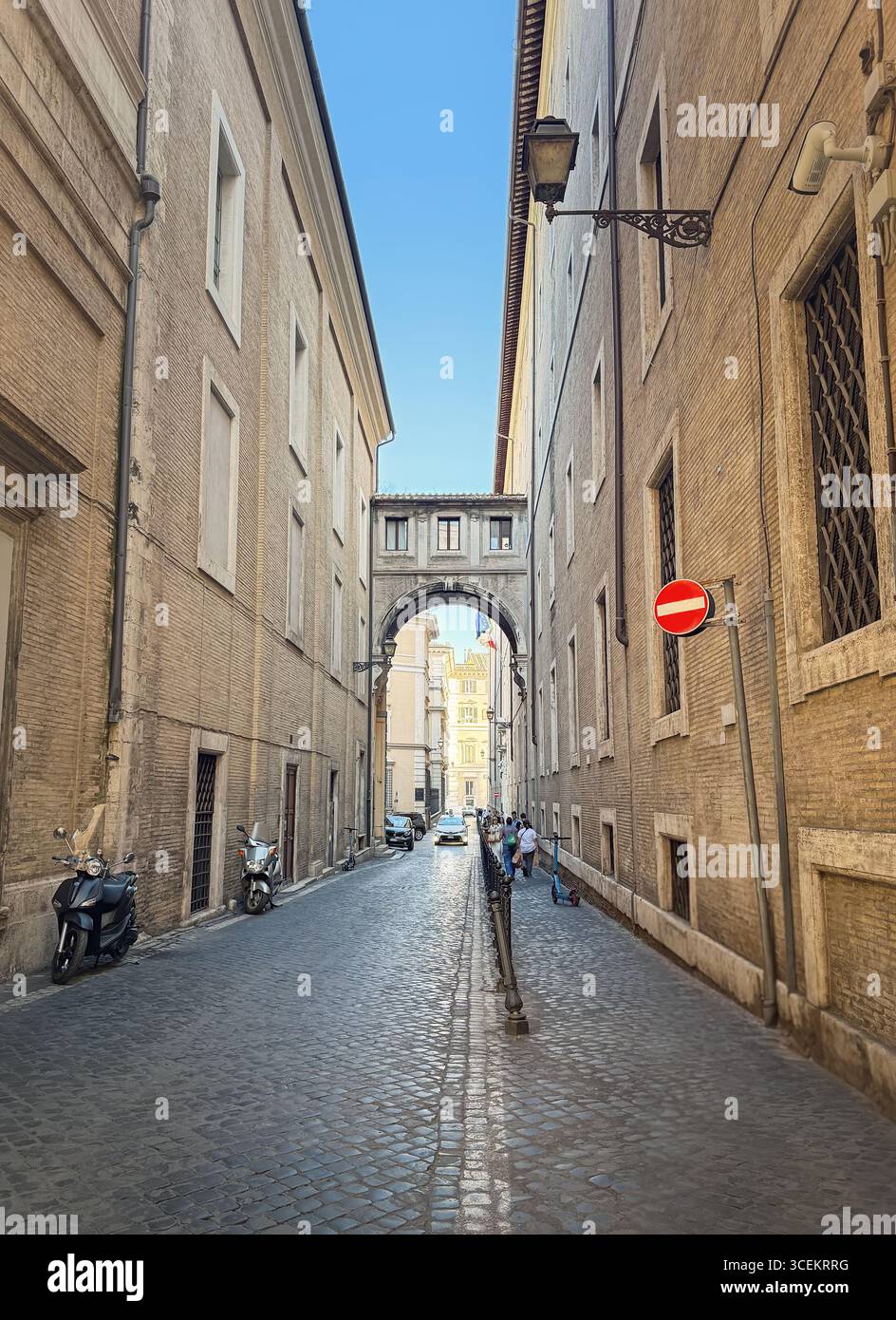 Eine lange, enge Kopfsteinpflasterstraße, gesäumt von hohen Backsteinhäusern und einem einzigartigen Bogengang in Rom, Italien. Geparkte Motorroller und ein paar Leute laufen Stockfoto