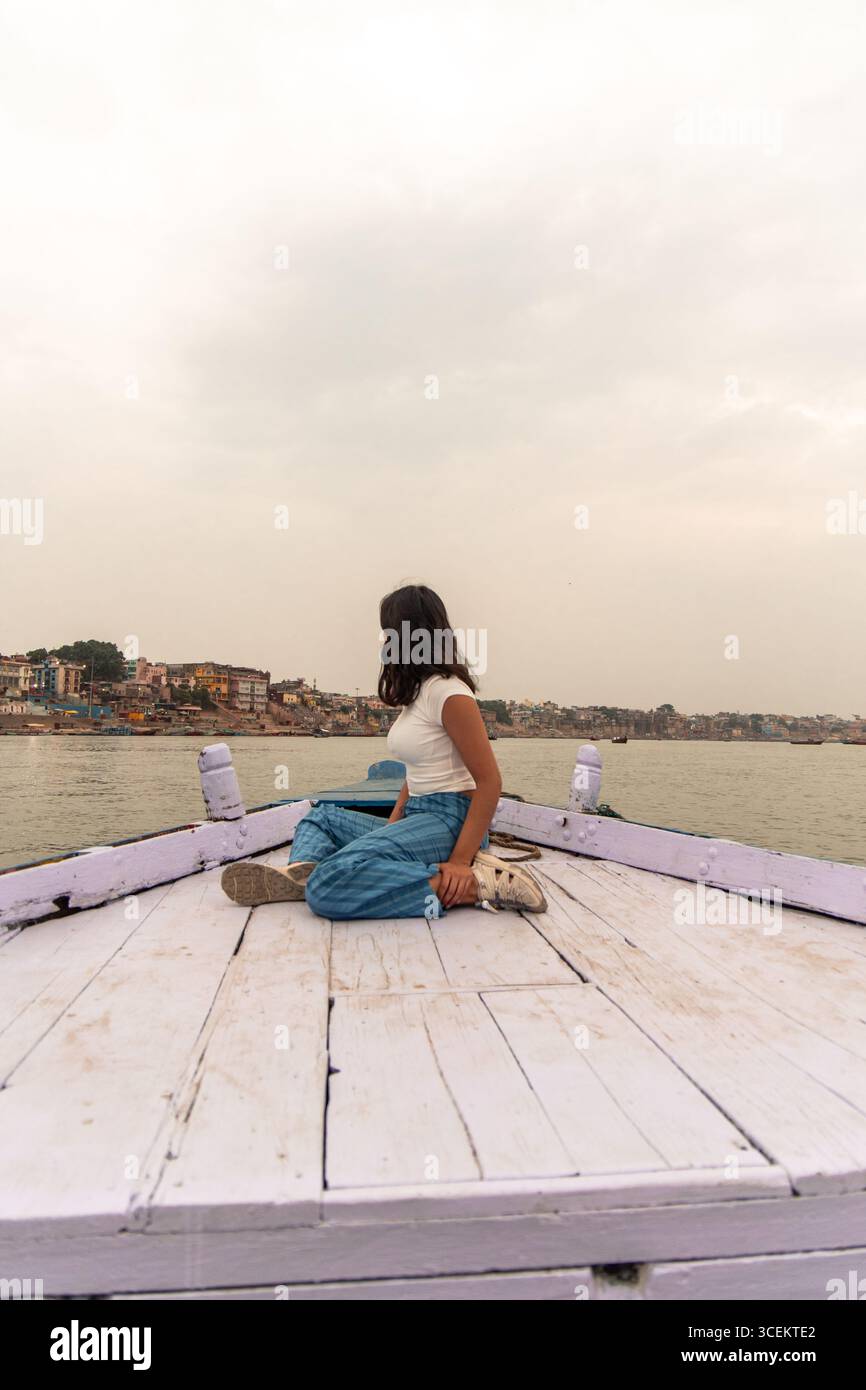 Junge Frau, die auf einem Holzboot auf dem Ganges-Fluss sitzt, die Stadtansicht von Varanasi mit bedecktem Himmel im Hintergrund bewundern und authentische Ind. Erleben Stockfoto