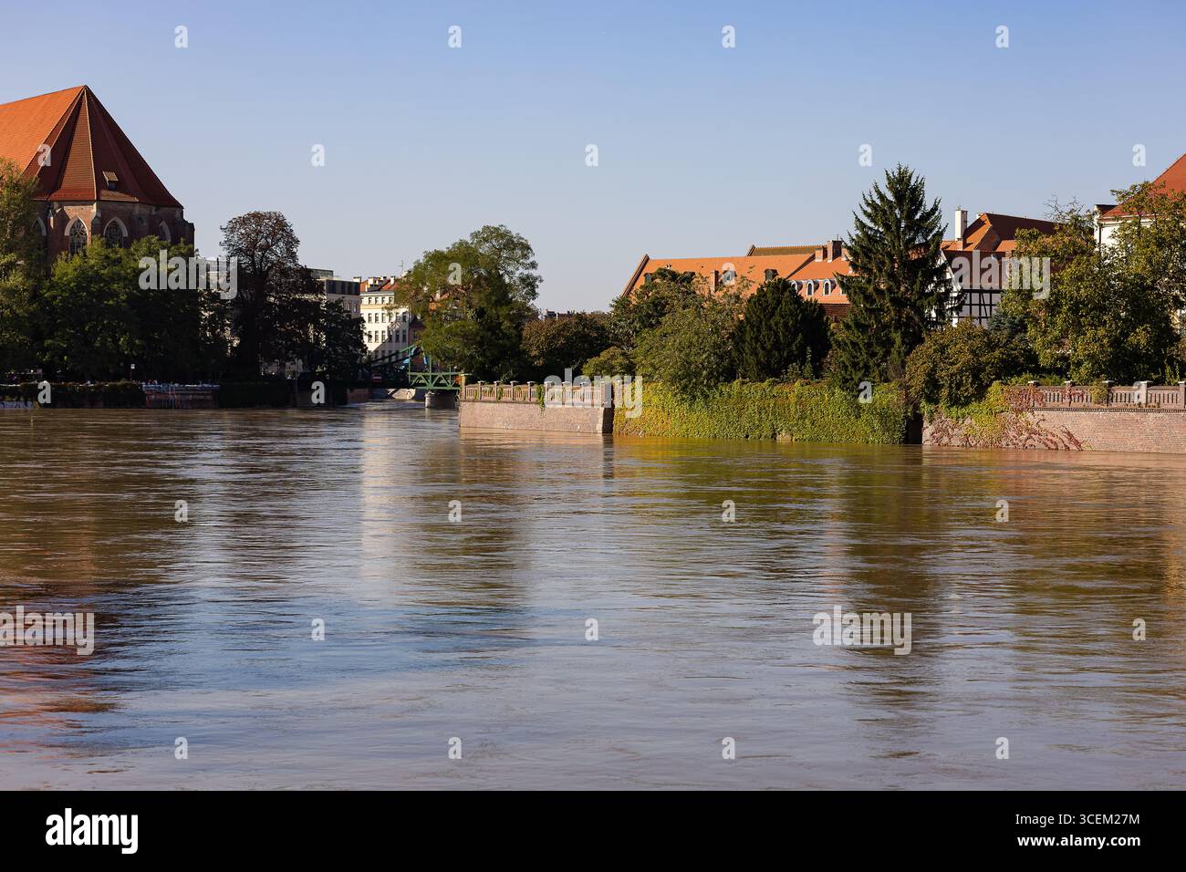 Breiter städtischer Fluss mit starker Strömung nach starken Bergregnern, reflektierenden Häusern und Bäumen unter klarem Himmel. Naturgewalt und Hochwasserrisiko Stockfoto