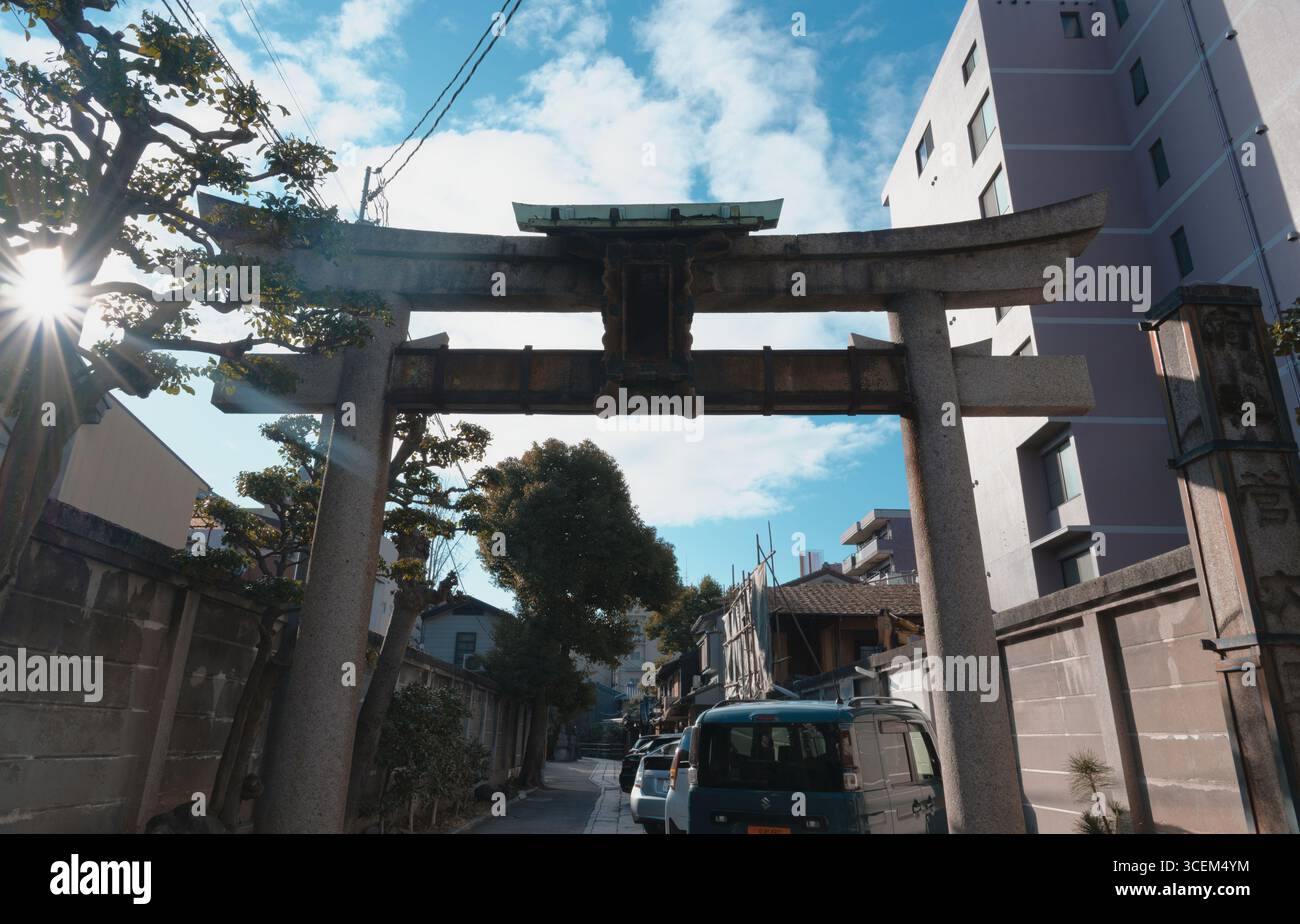 Morgensonnenlicht zieht durch die Bäume und beleuchtet ein verwittertes Torii-Tor in einer ruhigen Kyoto-Straße Stockfoto
