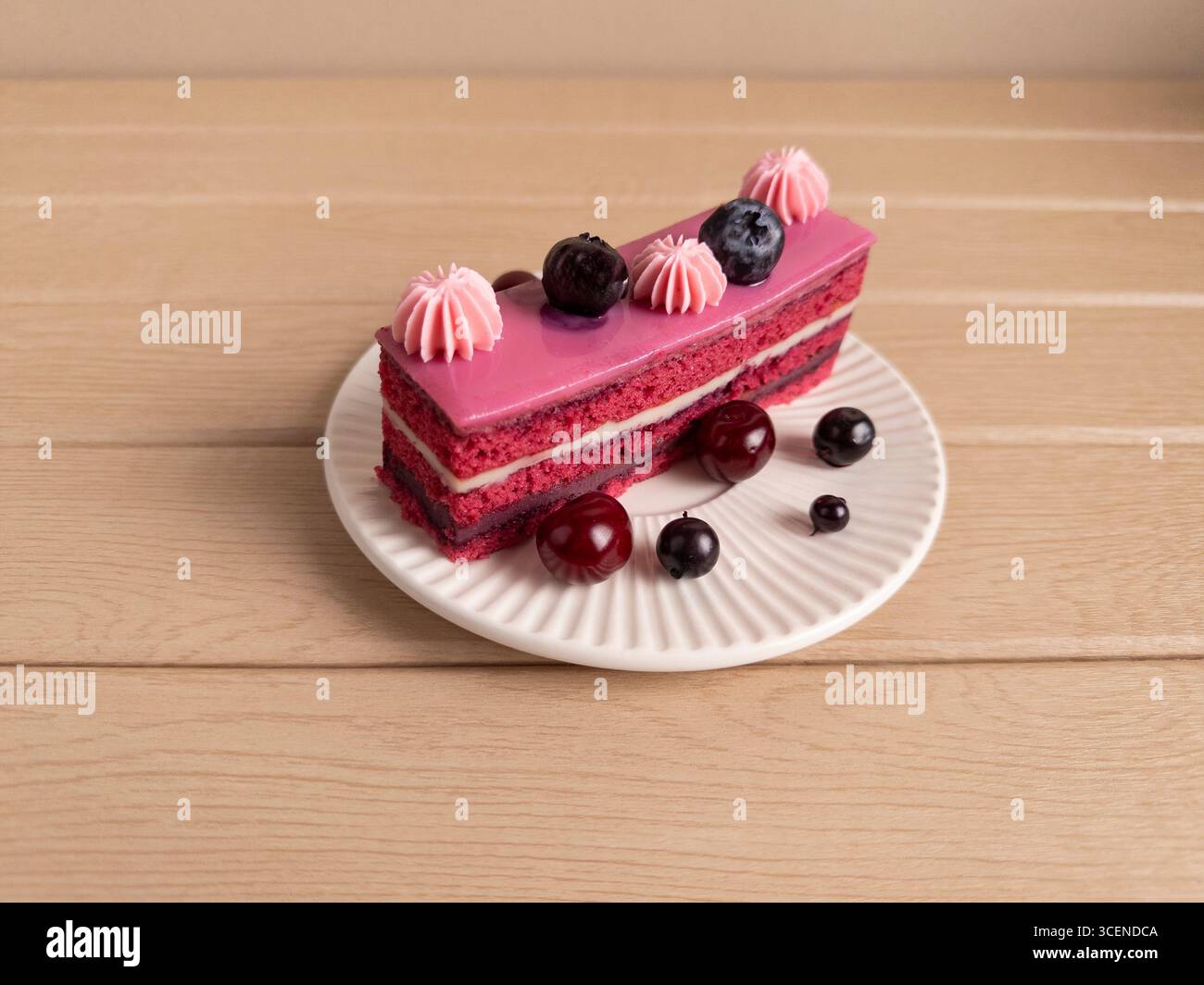 Leckere Scheibe rosafarbener Beerenkuchen mit Sahne, garniert mit Heidelbeeren und Kirschen, serviert auf einem weißen Teller. Ein perfektes Lebensmittelbild für die Werbung für Bäckerei Stockfoto